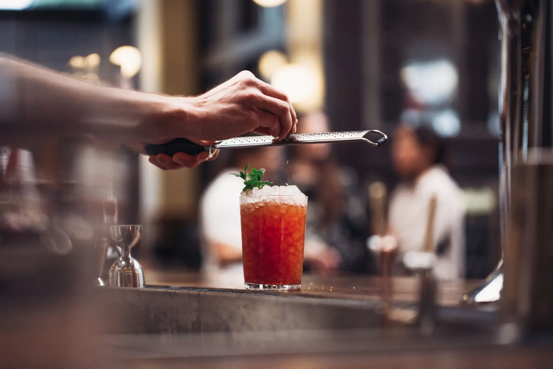 fruit cocktail on table topped with ice while a bartender grates ingredient over glass