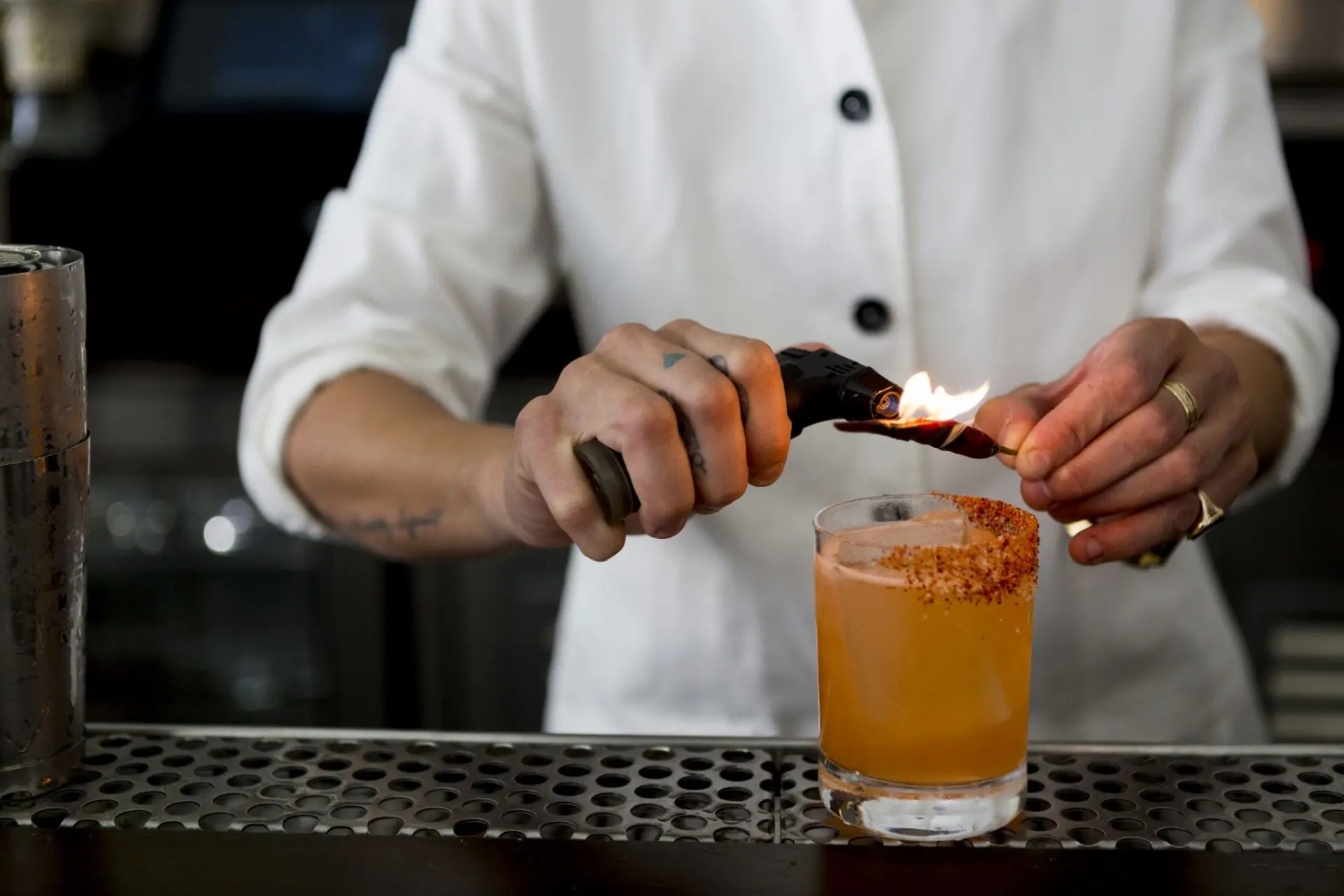 cocktail with chilli powder on the rim while bartender roasts chilli pepper with flamethrower