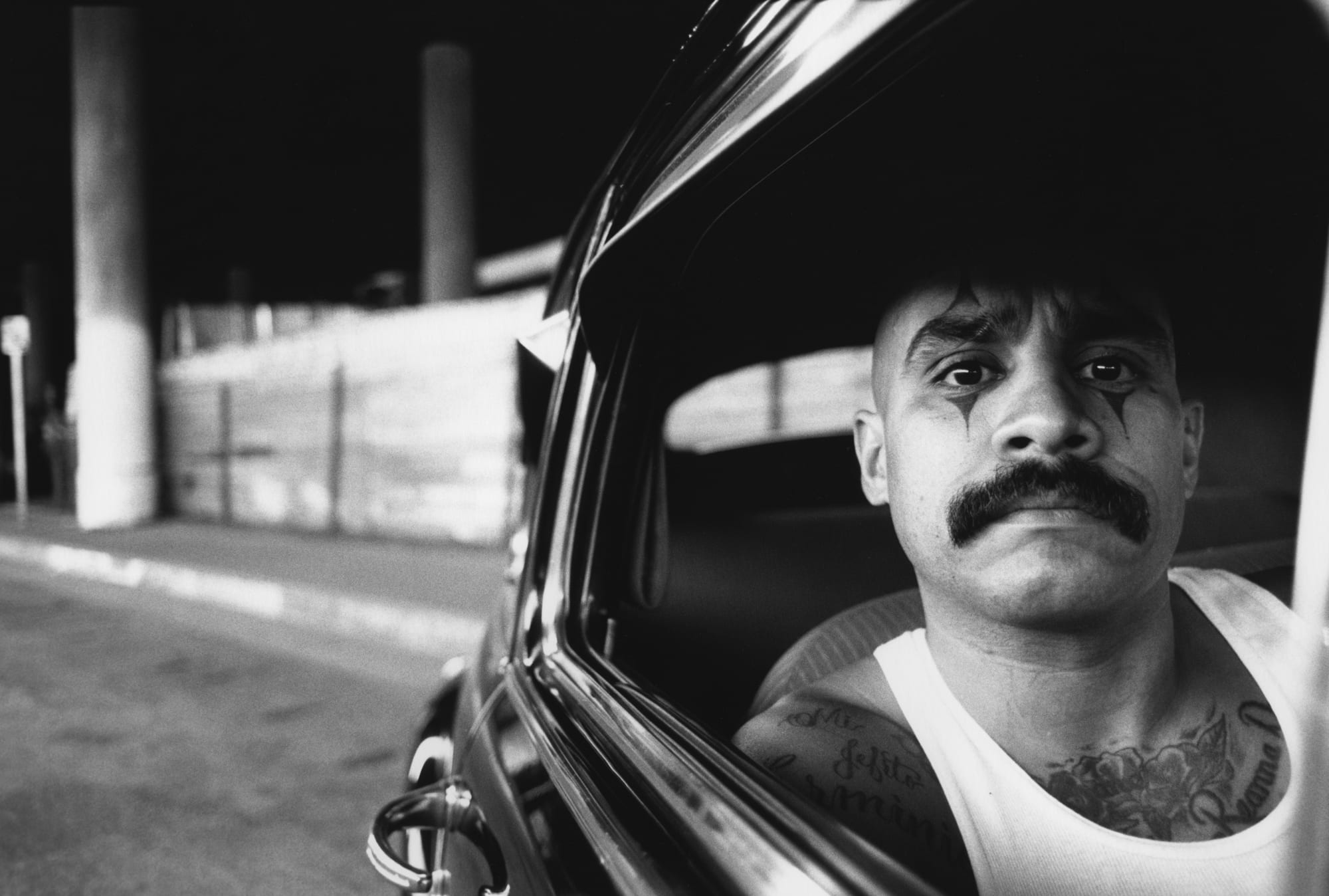 man with clown tattoos around eyes sitting on a car, looking out a window