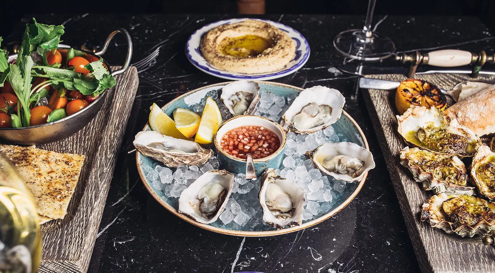 a table with plates of raw oysters, grilled oysters and hummus