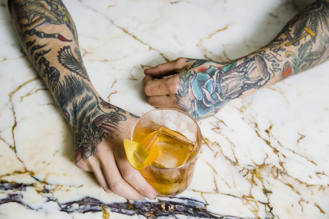 person with tattoo sleeves holding a cocktail with hands on marble table
