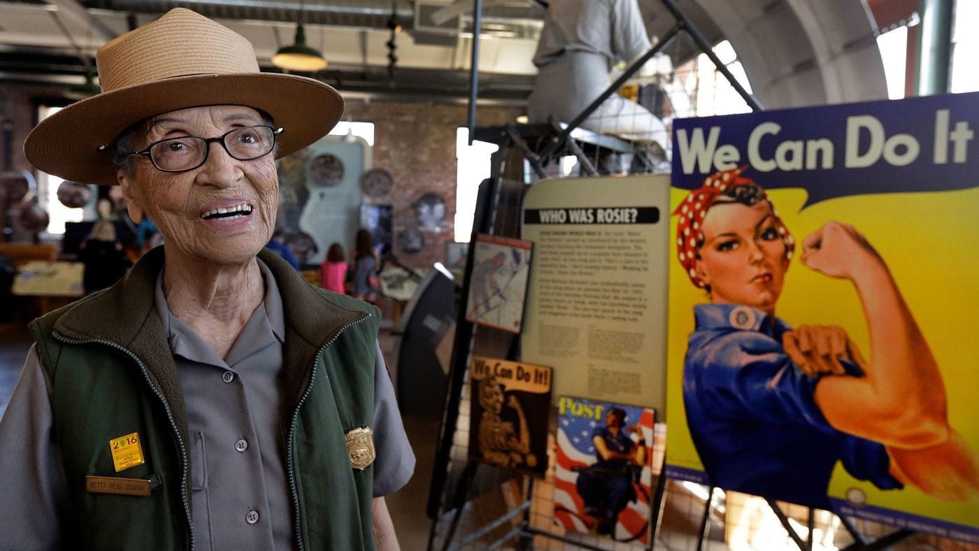 older woman smiling wearing forest ranger uniform next to rosie the riveter poster