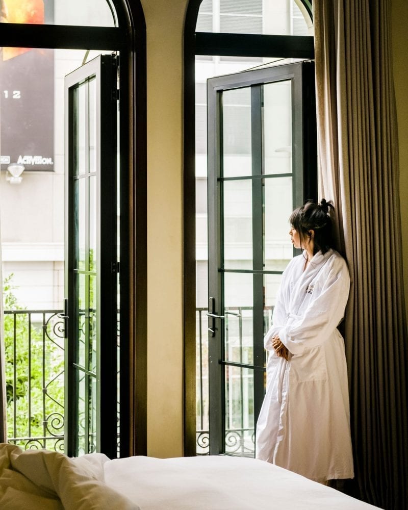 woman wearing bathrobe, looking outside hotel room from balcony door