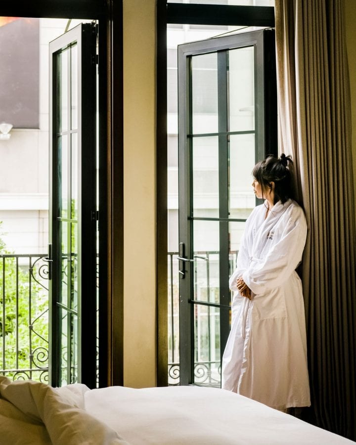 woman wearing bathrobe, looking outside hotel room from balcony door